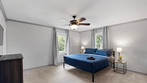 Une chambre à coucher comprenant un lit, deux tables de chevet, un ventilateur de plafond et une fenêtre avec des rideaux.