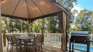 Une terrasse extérieure couverte, avec une table et des chaises, un barbecue, et une piscine en arrière-plan.