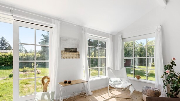 A bright, spacious room with large windows, a wooden floor, a wicker chair, a white chair, a small table, and a brown sofa. - Nice home in Jægerspris with kitchen (Jægerspris)