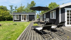 A wooden deck with lounge chairs and an umbrella, a small house with a porch, and a garden area. - Nice home in Jægerspris with kitchen (Jægerspris)