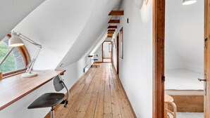 Un estrecho pasillo con suelo de madera, techo inclinado, un escritorio con lámpara y una silla.
