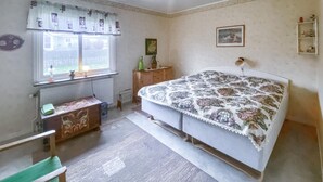Un dormitorio con cama, cómoda, radiador, ventana con cortinas y estante con figuritas.