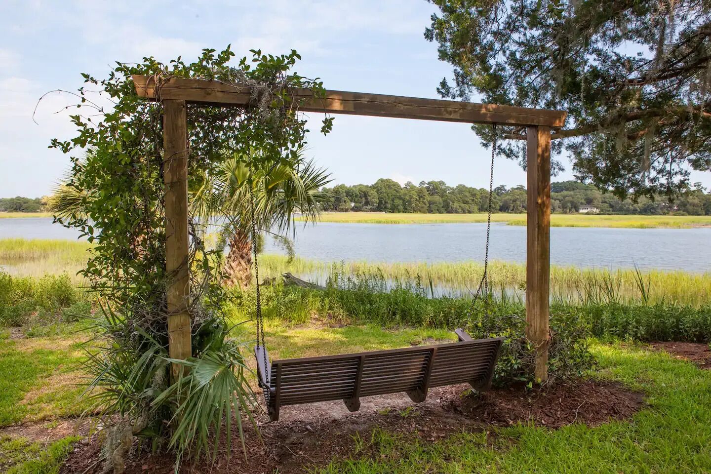 A wooden swing set with a bench, surrounded by greenery and a lake view.