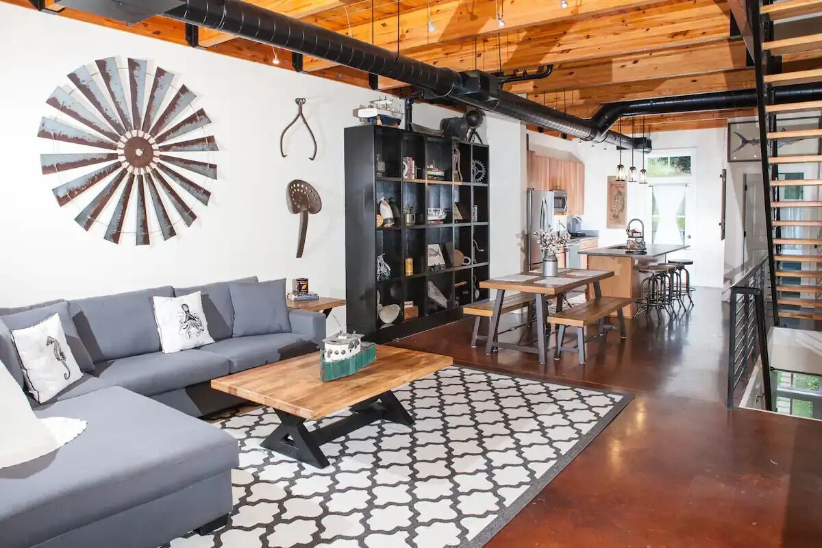 A modern living room with a gray sofa, wooden coffee table, and a patterned rug. A black bookshelf and a decorative fan are also visible.