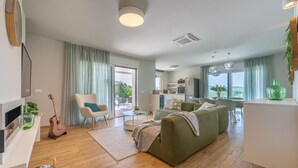 En moderne stue med en grøn sofa, et træbord og en guitar. En skydedør fører ud til en balkon med udsigt over grønne områder.