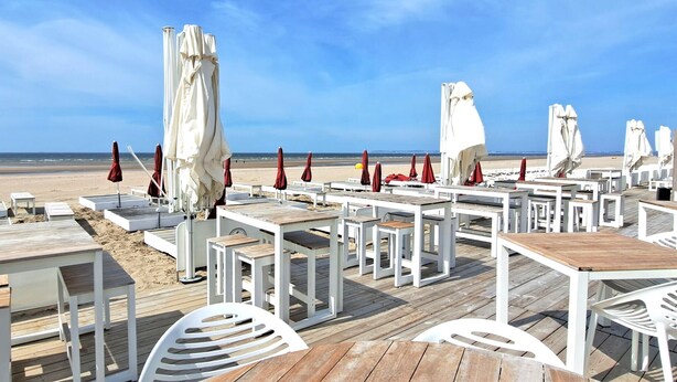 Un restaurante frente a la playa con mesas al aire libre, manteles blancos y sombrillas rojas.