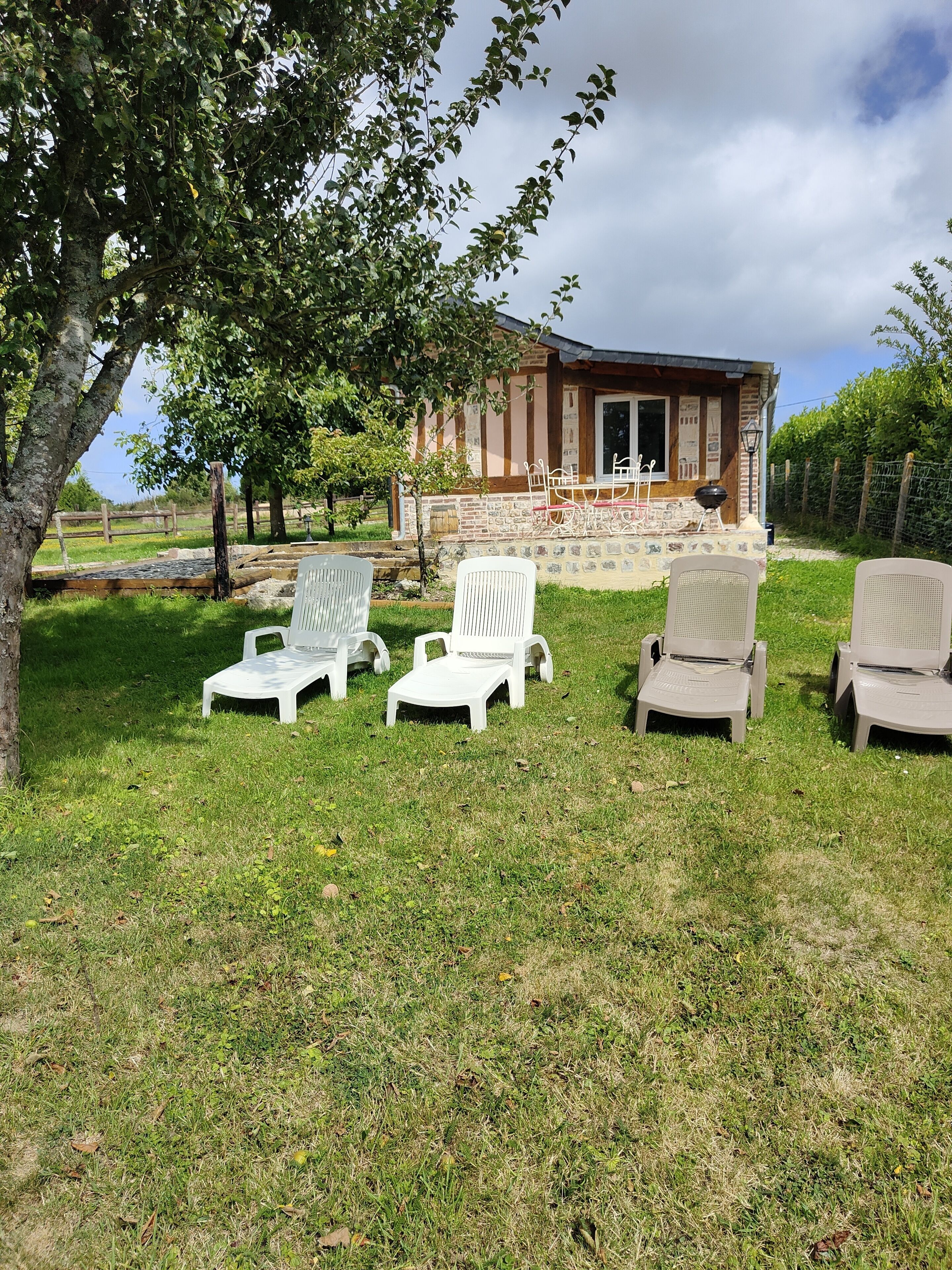 Un espace extérieur aménagé dans un style rustique, avec des fauteuils de jardin blancs et beiges, un petit bâtiment à la façade en briques et un arbre au premier plan.