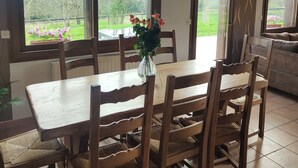 Een eethoek met een houten tafel en stoelen, grote ramen met uitzicht op groen, en een vaas met bloemen op de tafel.