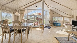 Ein geräumiger Raum mit hohen Decken, Holzböden, einem Essbereich mit Stühlen und Tisch, einer Sitzecke mit Sofa und Fernseher sowie großen Fenstern mit Blick ins Freie.