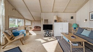 Ein gemütliches Blockhaus-Interieur mit Holzbalkendecke, einem Fernseher, einer Couch, einem Esstisch und einem Bücherregal.