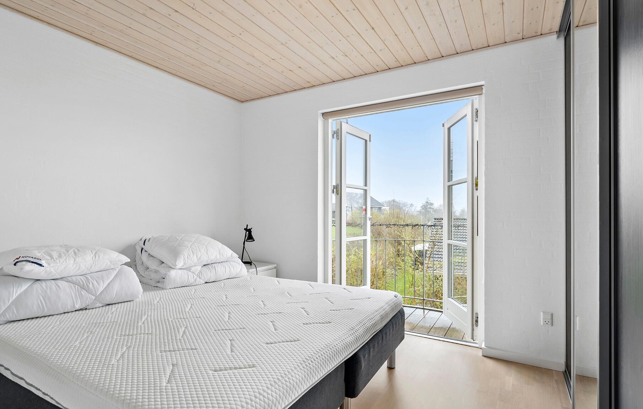 Ein Schlafzimmer mit einem Bett, einem Nachttisch, einer Lampe und einem großen Fenster mit Blick ins Freie.