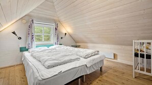 Ein gemütliches Schlafzimmer unter dem Dach mit schrägverlegtem Holzboden, einem großen Bett mit weißer Bettwäsche, einem Fenster mit Vorhängen und einem kleinen Nachttisch.