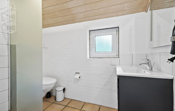 Een compacte badkamer met een wit toilet, een klein raam, een wastafel met spiegel en een prullenbak.