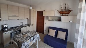 A compact kitchen with a gas stove, sink, and cabinets. There is a dining table with a patterned tablecloth. A blue sofa with two pillows is against the wall. A ship model is displayed above the sofa. - Park Snail - Alghero Loc. Caragol   (Alghero)