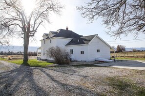 A two-story white house with a black roof, a gravel driveway, and a small garden area. - 1895 Luxury Farmhouse - Blodgett Canyon View -Sleeps 13 (Hamilton)