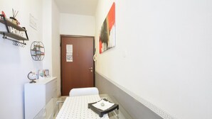 Un couloir étroit avec une table en verre, un meuble blanc à tiroirs, une porte en bois et un tableau encadré accroché au mur.