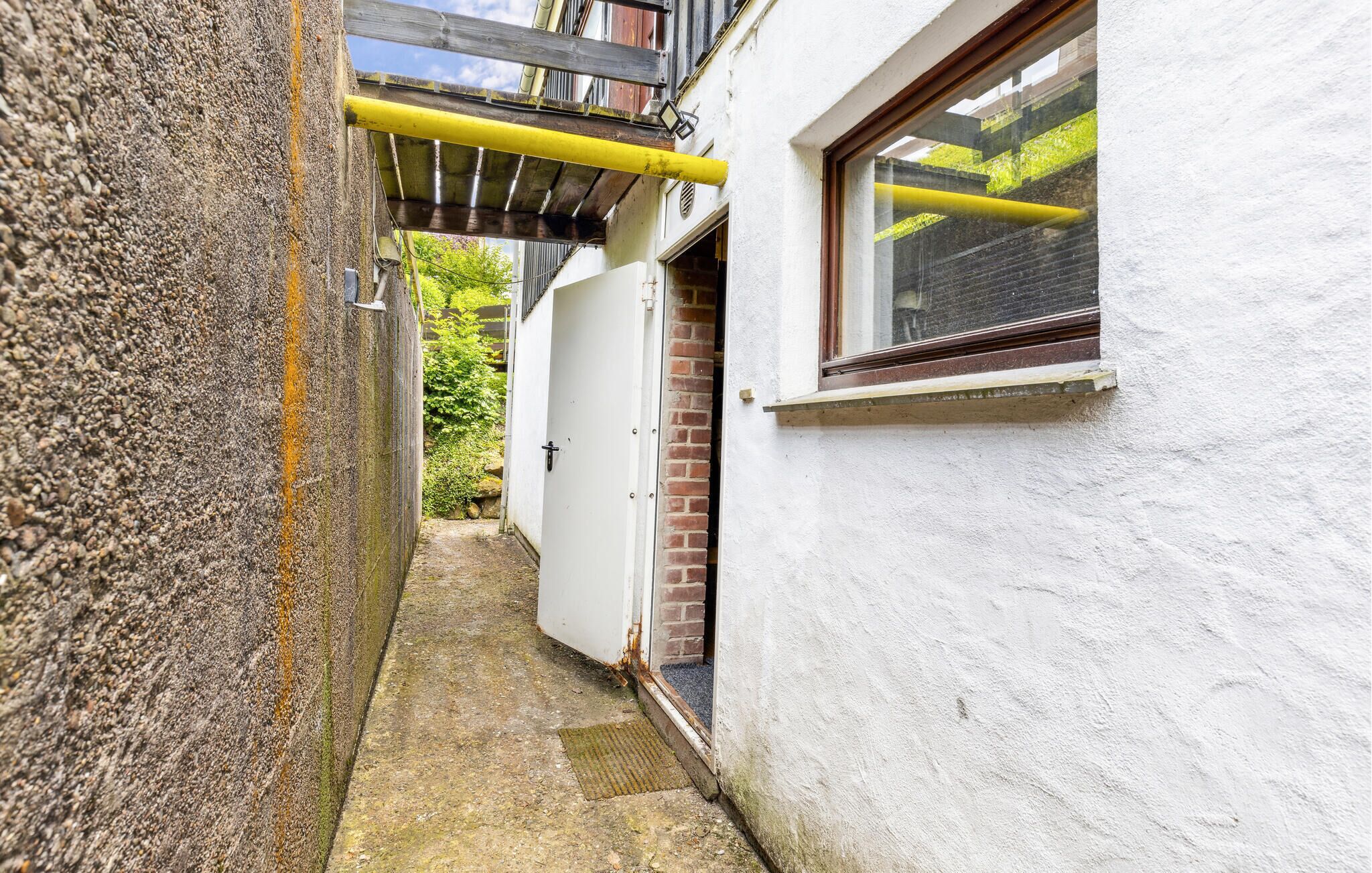 A narrow alleyway with a white building on one side and a wooden structure on the other. A yellow pipe runs along the top of the alley.
