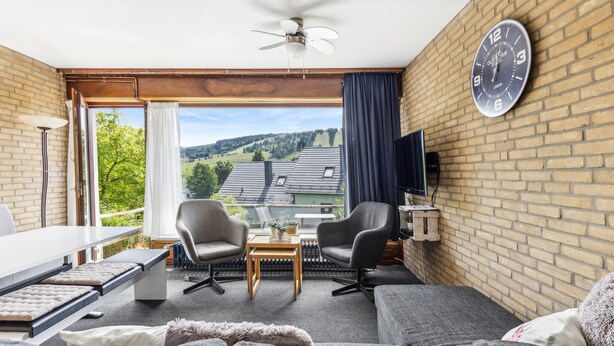 Un salon moderne comprenant un canapé, deux fauteuils, une table basse et un téléviseur fixé au mur en briques.