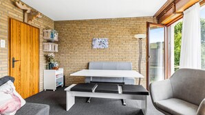 Une pièce avec un mur en briques, une porte en bois, un banc blanc, un canapé gris et une fenêtre donnant sur un paysage verdoyant.