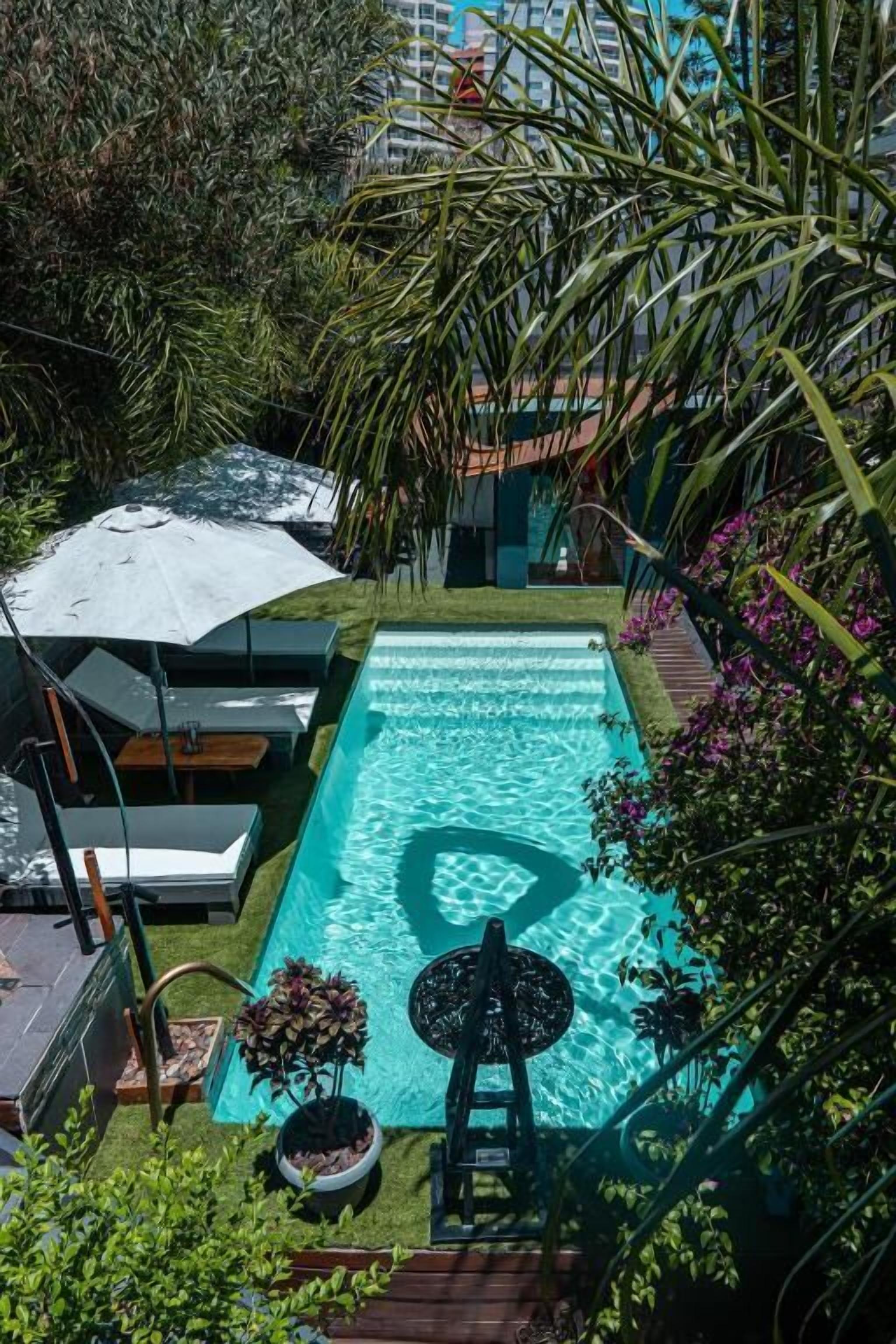 A rectangular swimming pool with a ladder and a small table with a chair, surrounded by lush greenery and a white umbrella.