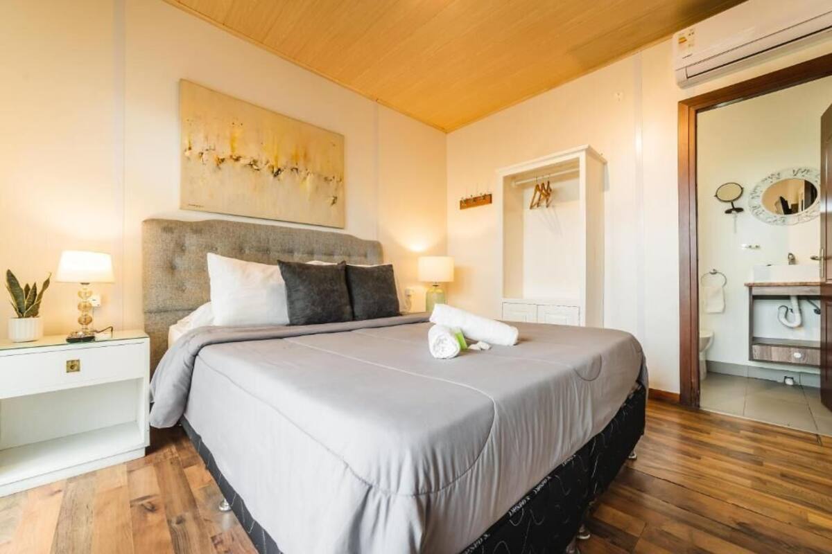 Un dormitorio con una cama grande, una mesita de noche con una lámpara, una obra de arte en la pared y un baño visible a través de una puerta abierta.