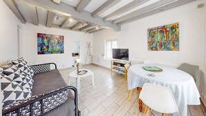 Een moderne woonkamer met zichtbare balken, een bank met gestreepte kussens, een eettafel met stoelen, en twee abstracte schilderijen aan de muur.