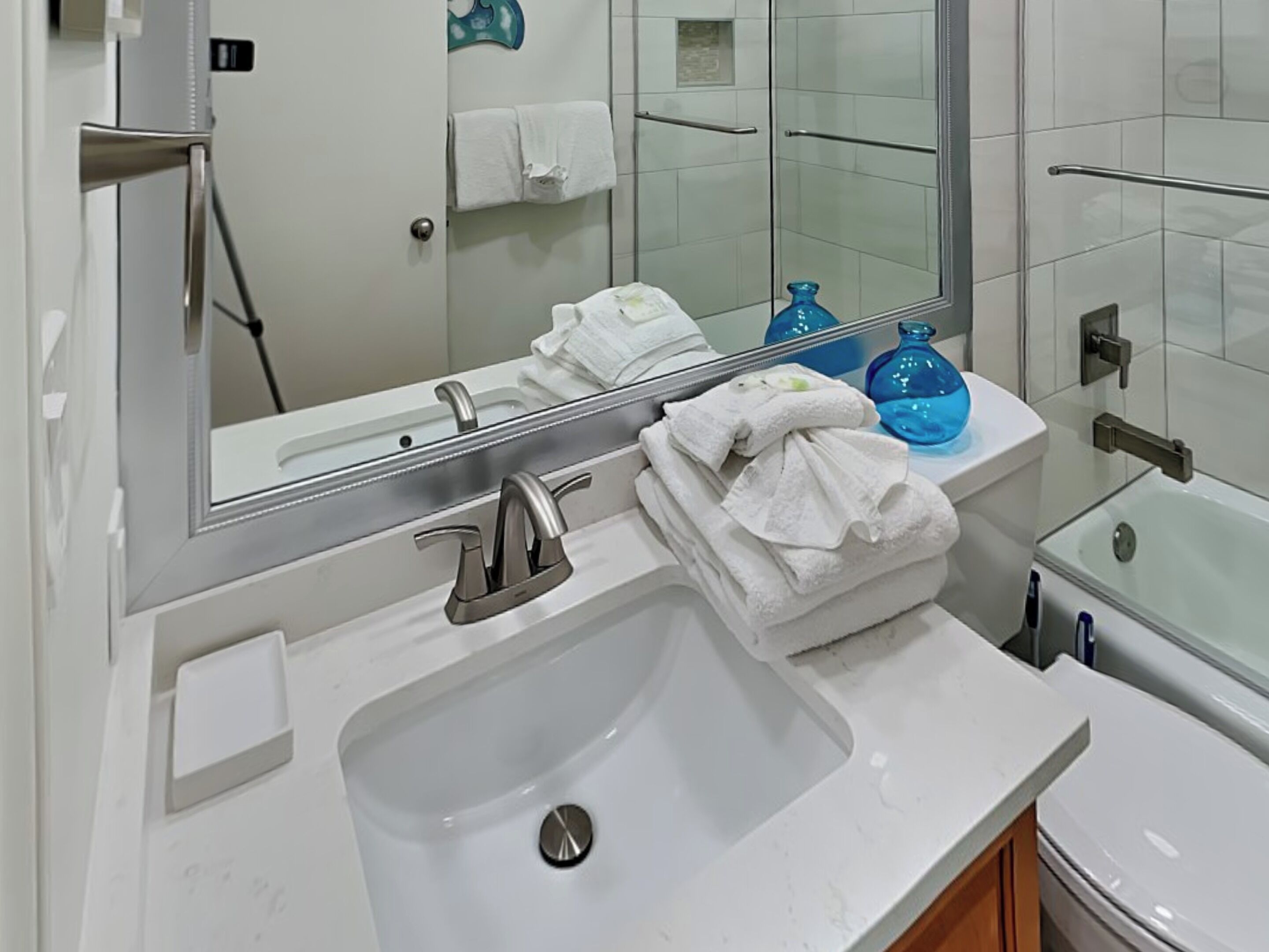 A bathroom with a white sink, a mirror, a glass shower enclosure, and a stack of white towels on a white shelf.