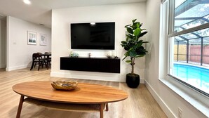 A modern living room with a flat-screen TV mounted on the wall, a dining table with chairs, a potted plant, and a view of a backyard swimming pool through a large window. - Sarasota modern house, heated saltwater pool, close to beaches and downtown! (Sarasota)