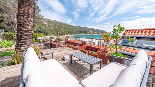 A rooftop terrace with outdoor seating, a palm tree, and a view of the sea and hills.