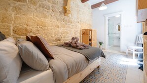 Quarto com parede de pedra, cama de madeira, cabideiro suspenso e banheiro visível por uma porta aberta.