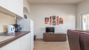Una cocina moderna con microondas, cafetera y refrigerador. Hay un mueble de madera con televisor de pantalla plana y una mesa pequeña con adornos. El cuarto tiene piso de madera y una puerta que conduce a otra área.