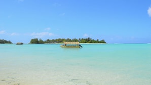 A boat named 'Captain Tamo's Cruiser' on clear turquoise water with a sandy beach and a small island with trees in the background. - Mangavai Bungalow (Rarotonga)
