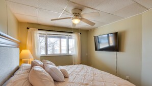 Une chambre à coucher avec un lit, un ventilateur de plafond, un téléviseur fixé au mur et une fenêtre avec des rideaux.