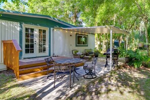 A covered patio with outdoor furniture, a grill, and a wooden deck leading to a house with a green roof and white exterior. - Cozy Fort Pierce Cottage on Farm Animal Rescue! (Fort Pierce)
