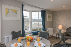 A dining area with a table set for breakfast, a view of the sea through large windows, and a stone wall. - Queens View (Dartmouth)