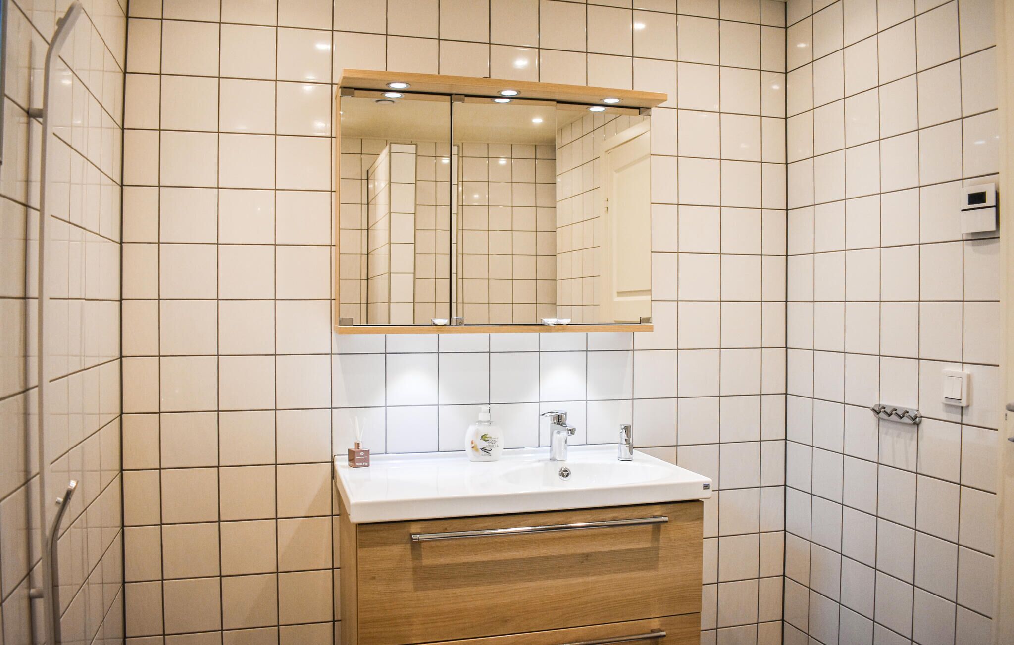 A modern bathroom with white tiles, a wooden vanity, a mirror, and a sink.