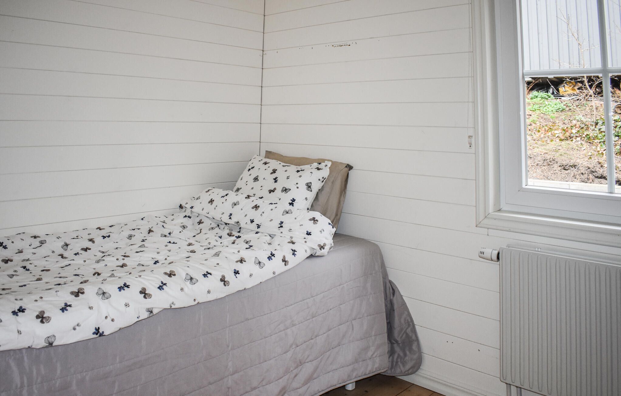 A single bed with a floral-patterned bedspread and pillows, a window with a view of greenery, and a radiator on the wall.