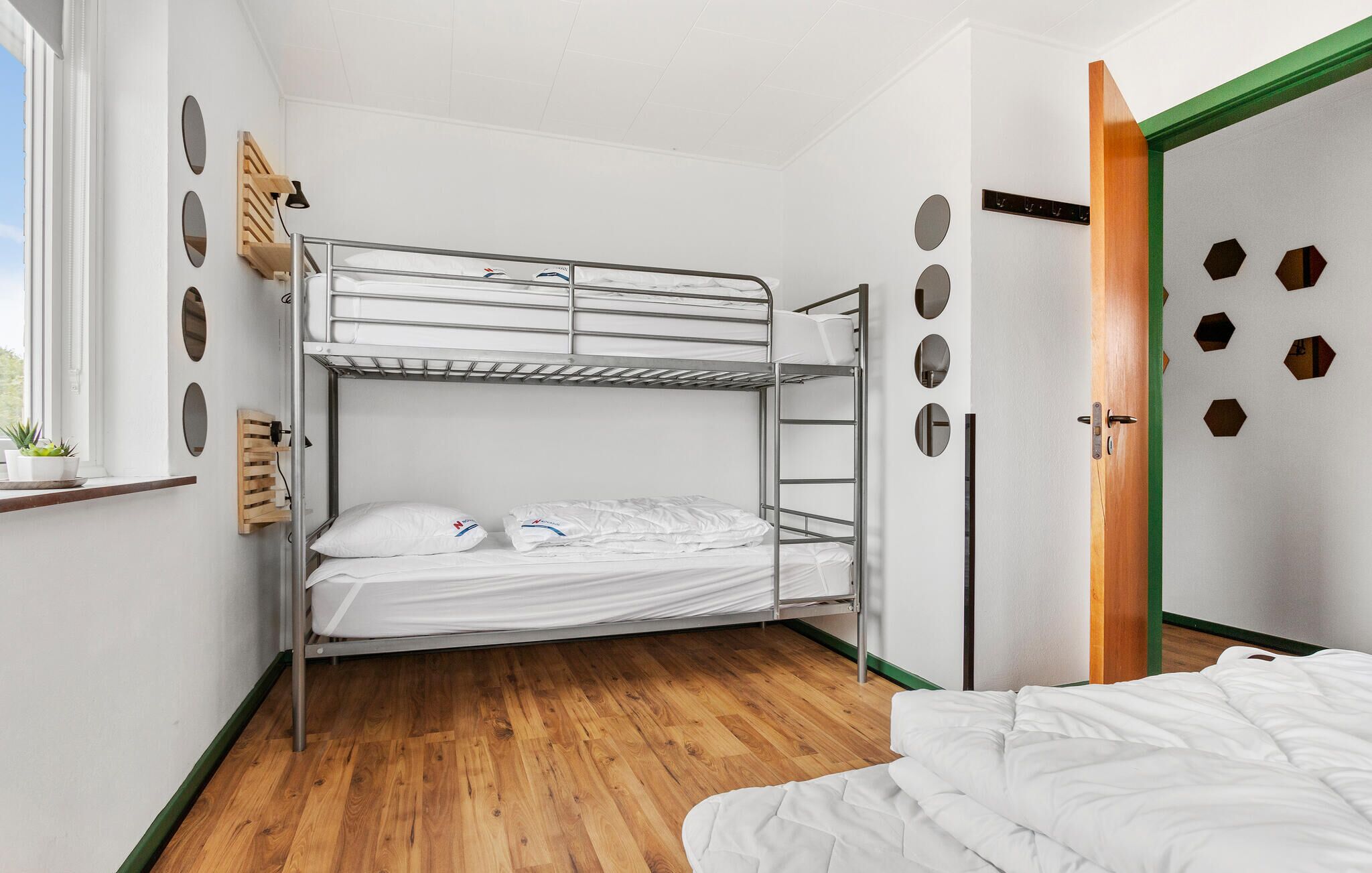 A bunk bed room with a window, a wooden shelf, and a green door with circular cutouts.