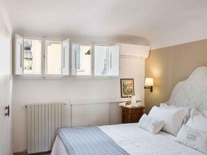 Een slaapkamer met een bed, nachtkastje, wandlamp, radiator en een raam met openslaande luiken.