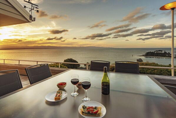 Une table avec une bouteille de vin, deux verres de vin rouge et deux assiettes de nourriture, le tout placé sur un balcon donnant sur un coucher de soleil sur la mer.