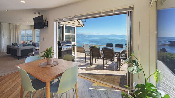 A modern living room with a wooden dining table, light-colored chairs, and a large window overlooking a scenic view.