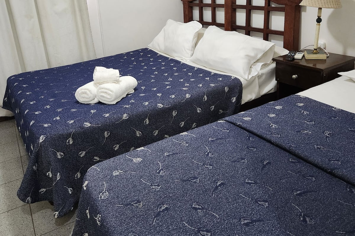Ein Hotelzimmer mit zwei Betten, jeweils mit blauen, gemusterten Bettdecken und weißen Kissen. Es gibt ein hölzernes Kopfteil, einen Nachttisch mit einer Lampe und eine Klimaanlage an der Wand.