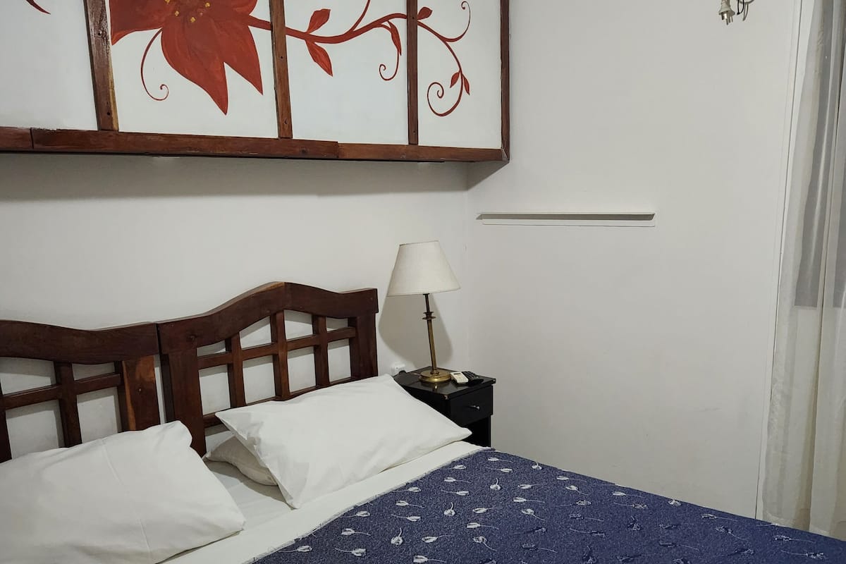 Ein ordentlich bezogenes Bett mit weißen Kissen und einer blumigen, blauen Tagesdecke, eine Nachttischlampe und eine wandmontierte Klimaanlage.