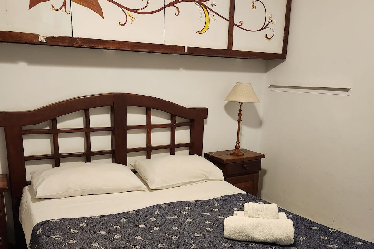 Ein ordentlich bezogenes Bett mit einem blumigen Überwurf, einem Holzkopfteil und einem Nachttischchen mit einer Lampe.