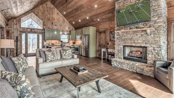 A spacious living room with a stone fireplace, a large window, and a kitchen area.