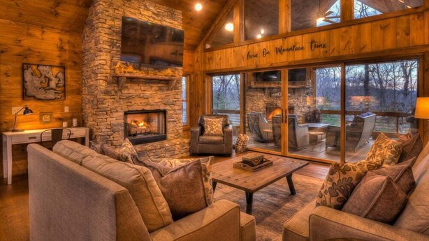 A cozy living room with a stone fireplace, comfortable seating, and large windows offering a view of the outdoors. - Eagles on the Ridge - Nearby River Access | Hot Tub | Game Room (Blue Ridge)
