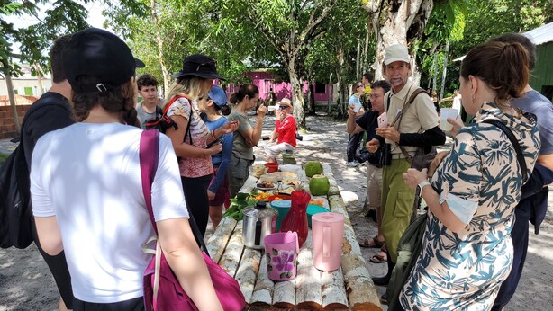 A group of people gathered around a table with food and drinks, standing in an outdoor area with trees and a rustic building in the background. - POUSADA CANTO DOS PÁSSAROS (Manaus)
