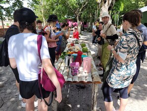 Un groupe de personnes s’est rassemblé autour d’une table où étaient disposés de la nourriture et des boissons, dans un espace extérieur agrémenté d’arbres et où l’on pouvait apercevoir, au loin, un bâtiment à l’allure rustique.