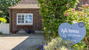 Een vakantiehuisje van baksteen met een wit raam, omringd door groen en een bord waarop staat 'Syl Homes SYLT COTTAGE 1'.