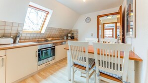 A kitchen with a window, a clock, and a dining area with white chairs. - Sylt Homes Auster 4 - Appartement Auster No.4 (Sylt)